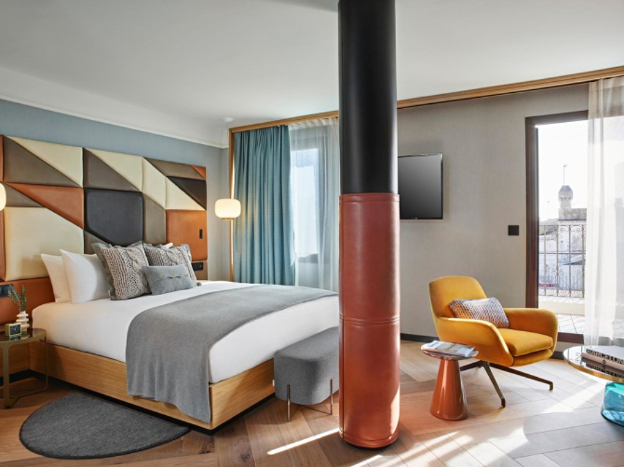 bedroom with geometric headboard, yellow armchair and orang side table, grey bench