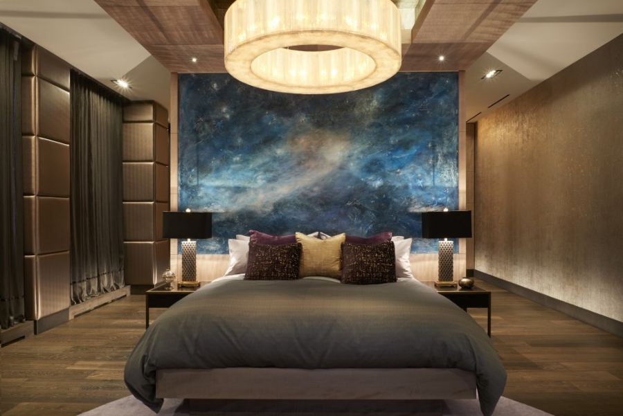 Modern Contemporary Interior Design by Fleur-de-lis Interior Design. This bedroom with a dark ambiance has a gold round chandelier, an enormous painting in blue as a bedframe, and two side tables with black lamps. 