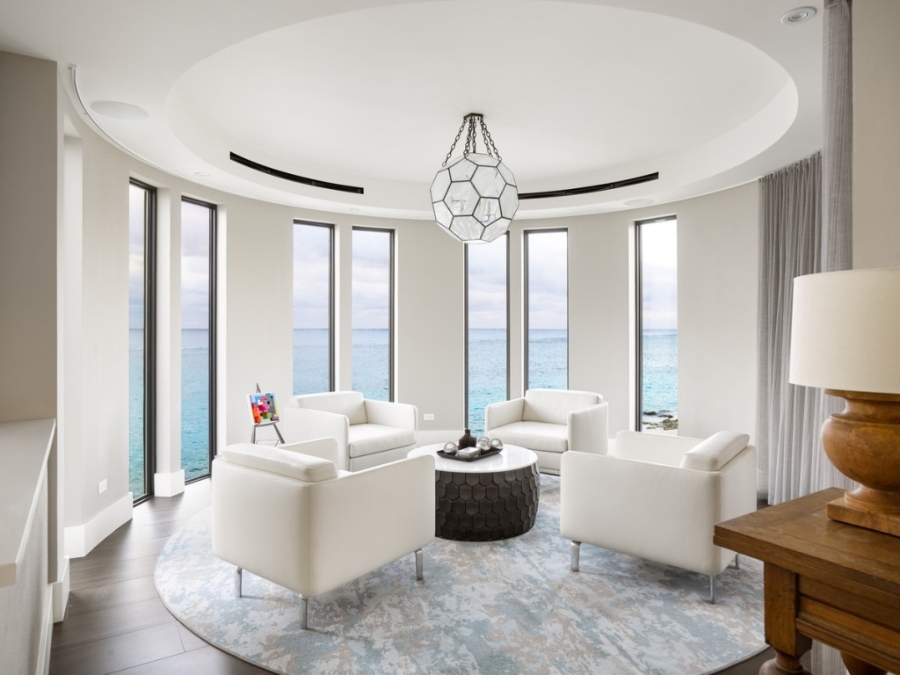 Modern Contemporary Interior Design by Fleur-de-lis Interior Design. This white seating area with a view of the ocean has four white armchairs and between these armchairs is a coffee table in brown with a white countertop, a round chandelier, and a round rug in neutral colors. 