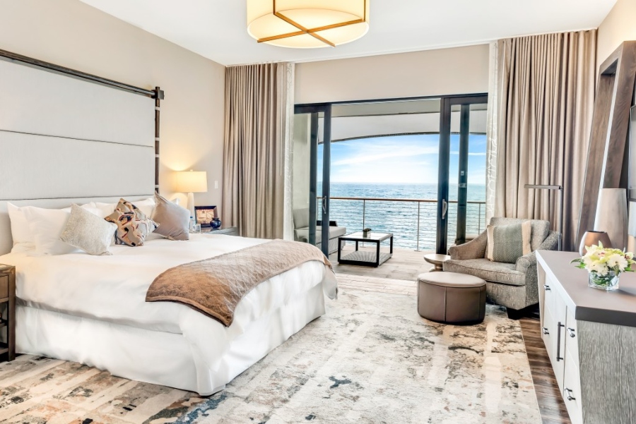 Modern Contemporary Interior Design by Fleur-de-lis Interior Design.This bedroom with a view of the ocean has neutral walls, a white bed frame, a grey armchair, a brown stool, and a rug in neutral. 