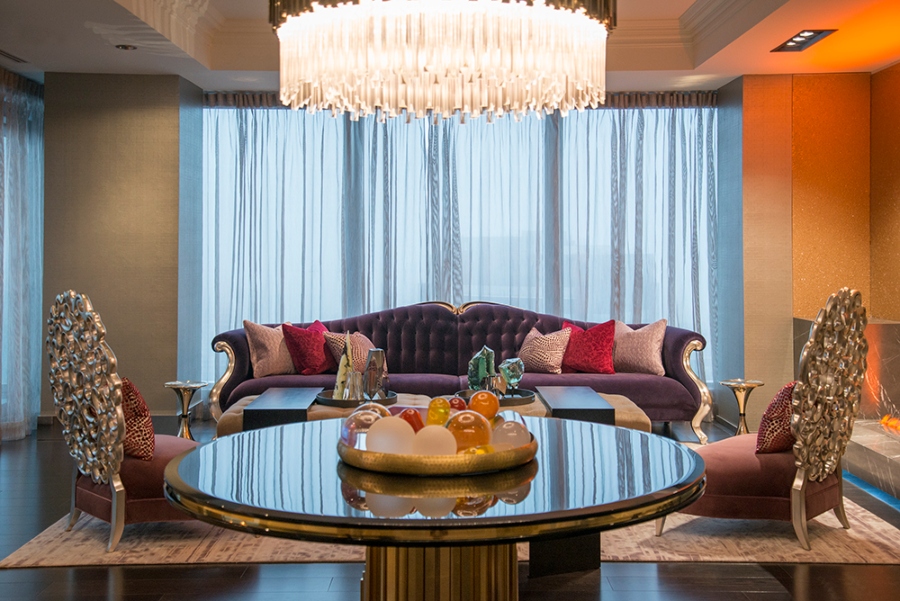 Modern Contemporary Interior Design by Fleur-de-lis Interior Design. This reception room with a dark ambiance and eclectic design has a purple sofa with gold details, two armchairs in purple with gold structure, a big chandelier in crystal, and a big round table in gold. 