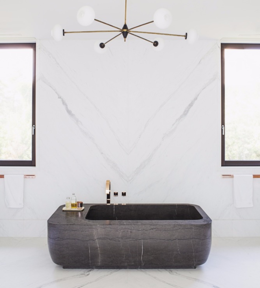The Best Interior Design Ideas By Audax. This bathroom with white marble walls and floor has a dark bathtub with golden feautures.